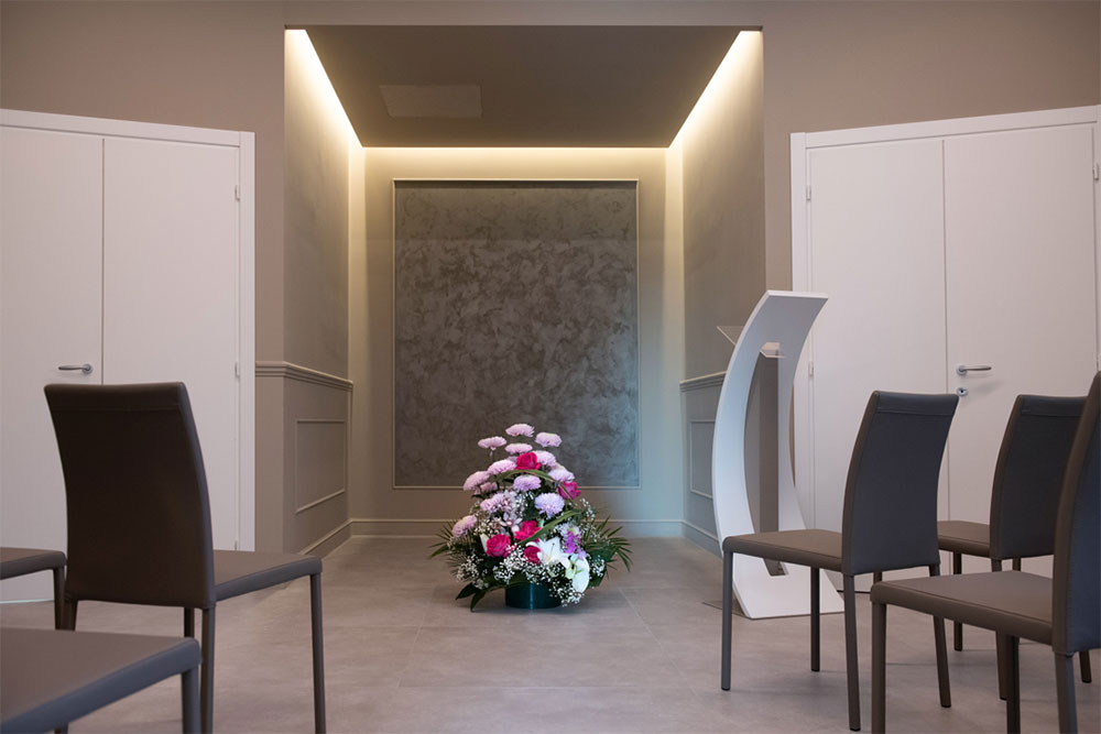 stanza allestita per veglia funebre con sedie, fiori e spazio dedicato per la bara illuminato
