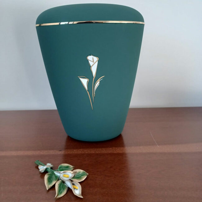 urna per ceneri con inciso un mazzo di calle bianche dallo sfondo color verde petrolio su un tavolo di legno